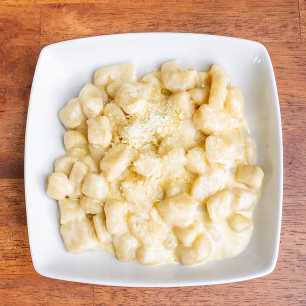 A white square bowl filled with creamy gnocchi topped with grated Parmesan cheese on a wooden table.