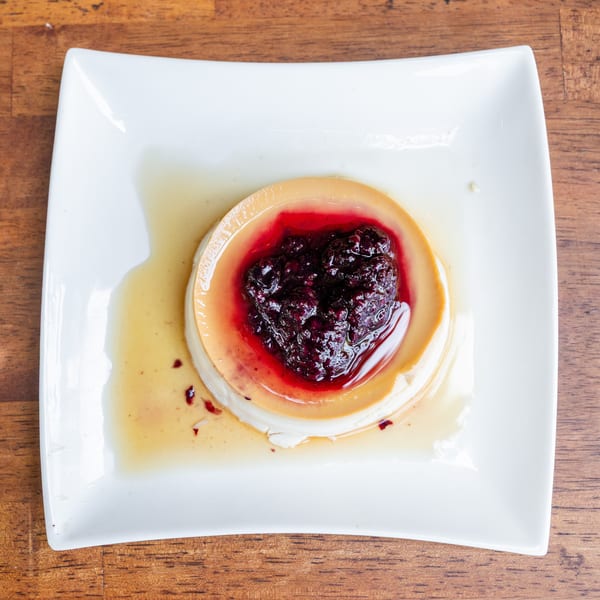 Flan dessert with a heart-shaped berry compote on a white square plate, on a wooden table.