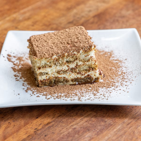 A slice of tiramisu with cocoa powder on a white rectangular plate, on a wooden table.