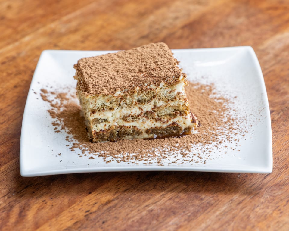 A slice of tiramisu with cocoa powder on a white rectangular plate, on a wooden table.