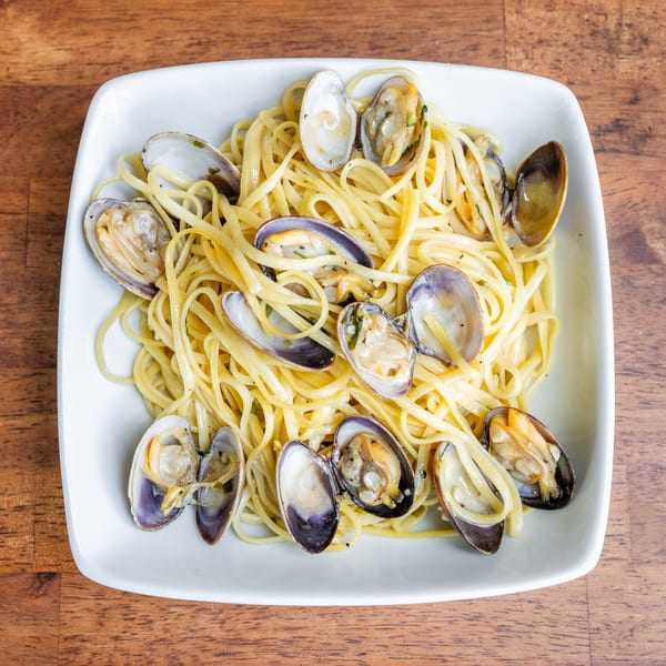 A white rectangular plate filled with linguine pasta and clams in a light sauce, on a wooden table.