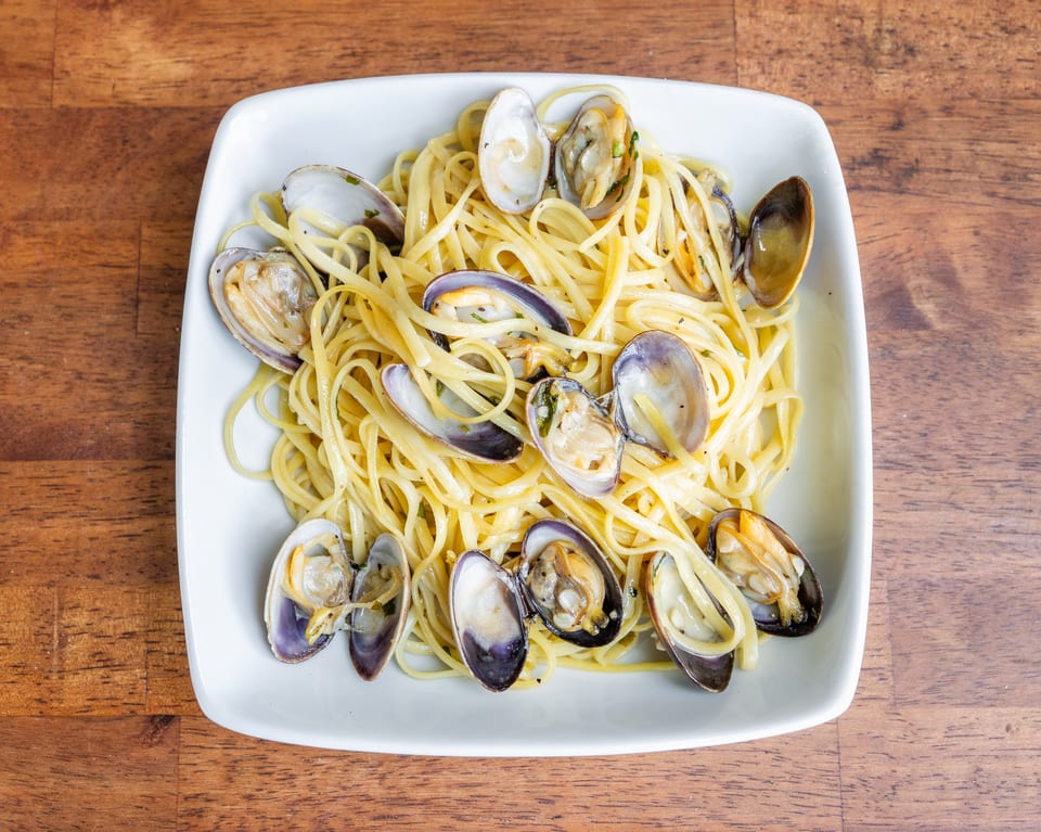 A white rectangular plate filled with linguine pasta and clams in a light sauce, on a wooden table.
