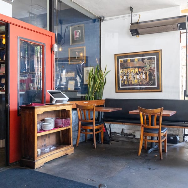 Restaurant interior with red door, wooden chairs, black booth seating, and a painting of a bustling street.
