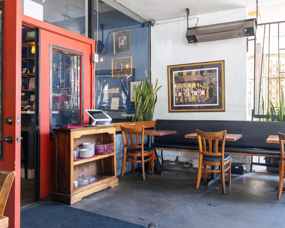 Restaurant interior with red door, wooden chairs, black booth seating, and a painting of a bustling street.