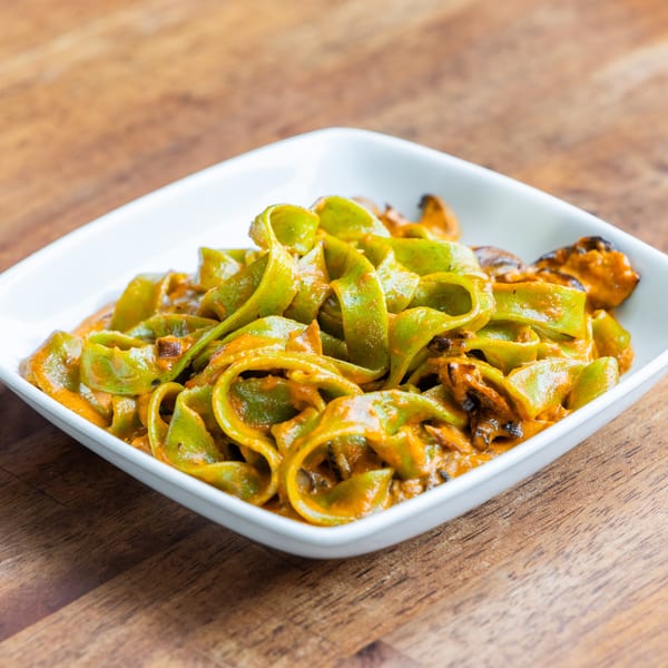Green pappardelle pasta with a rich orange sauce and mussels in a white bowl on a wooden table.