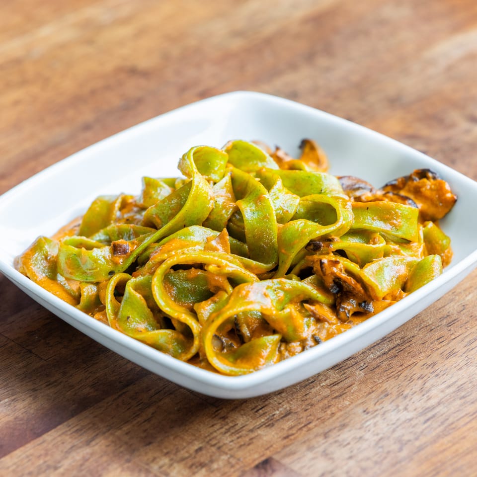 Green pappardelle pasta with a rich orange sauce and mussels in a white bowl on a wooden table.