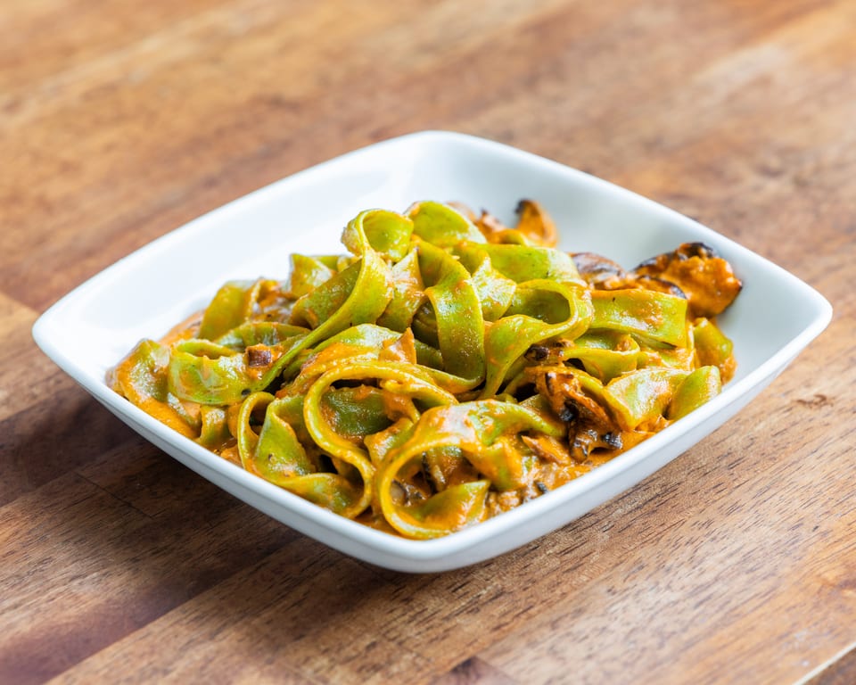 Green pappardelle pasta with a rich orange sauce and mussels in a white bowl on a wooden table.