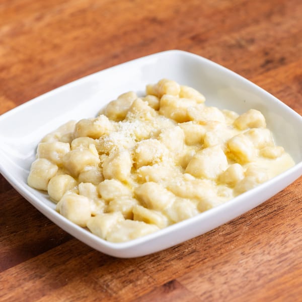 A white square bowl filled with creamy gnocchi topped with grated cheese on a wooden table.