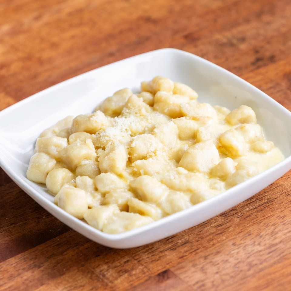 A white square bowl filled with creamy gnocchi topped with grated cheese on a wooden table.