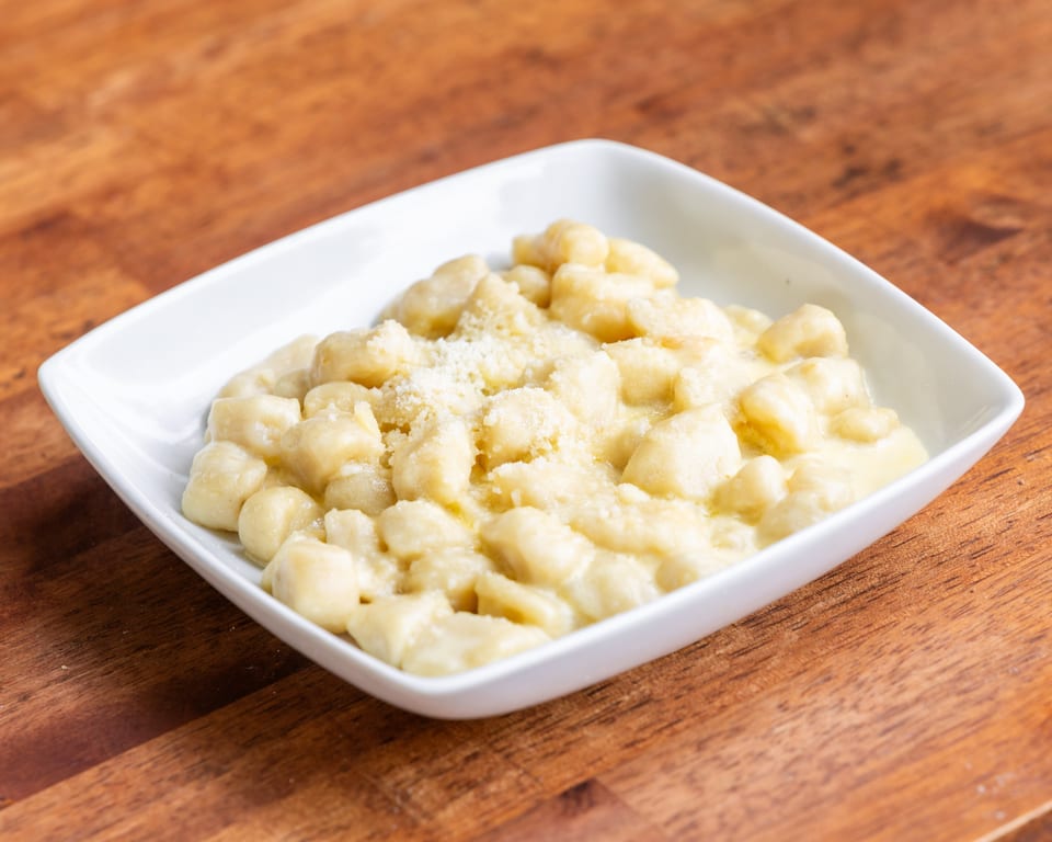A white square bowl filled with creamy gnocchi topped with grated cheese on a wooden table.