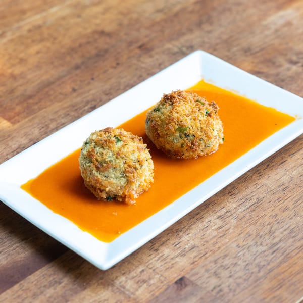 Two golden-brown arancini balls with herbs in a vibrant orange sauce on a white rectangular plate.