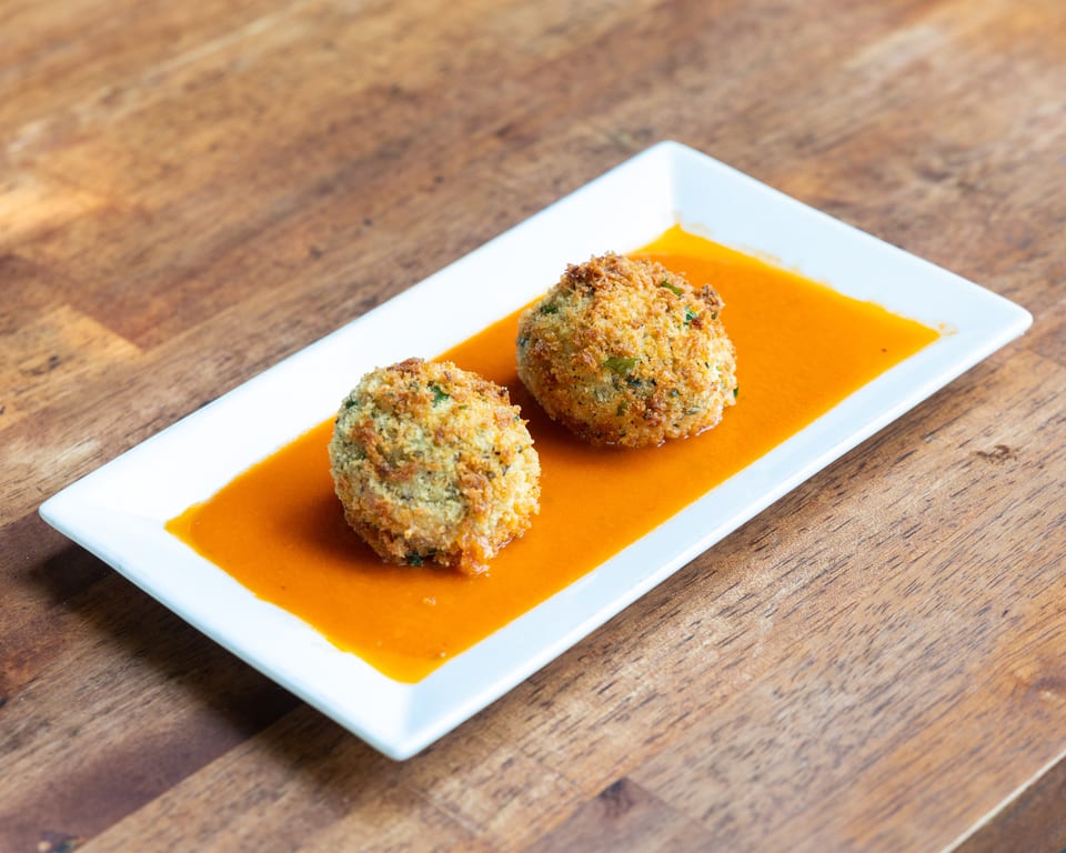 Two golden-brown arancini balls with herbs in a vibrant orange sauce on a white rectangular plate.