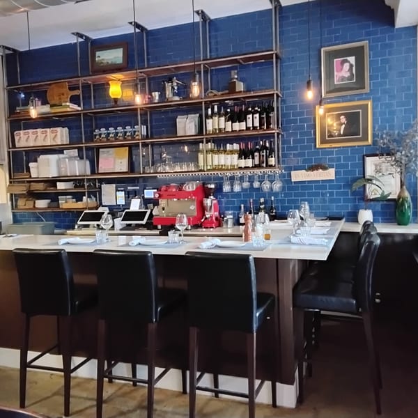 A restaurant bar with a blue tiled wall, shelves of wine, and black bar stools.