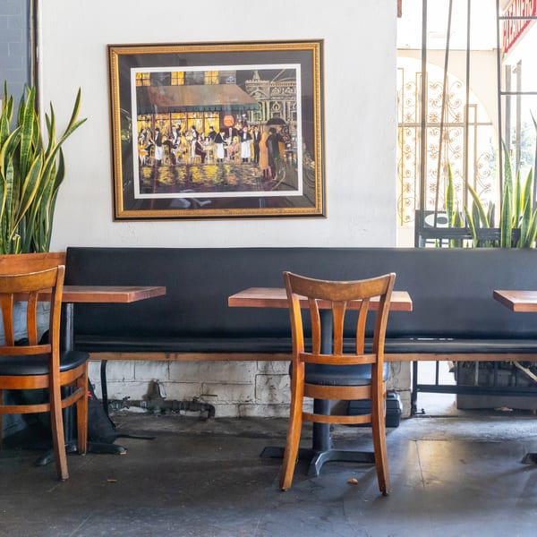 Restaurant interior with a long black booth, wooden tables and chairs, and a framed painting.