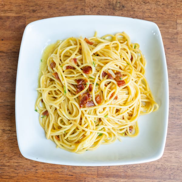 Spaghetti carbonara with crispy bacon bits and fresh herbs on a white square plate.