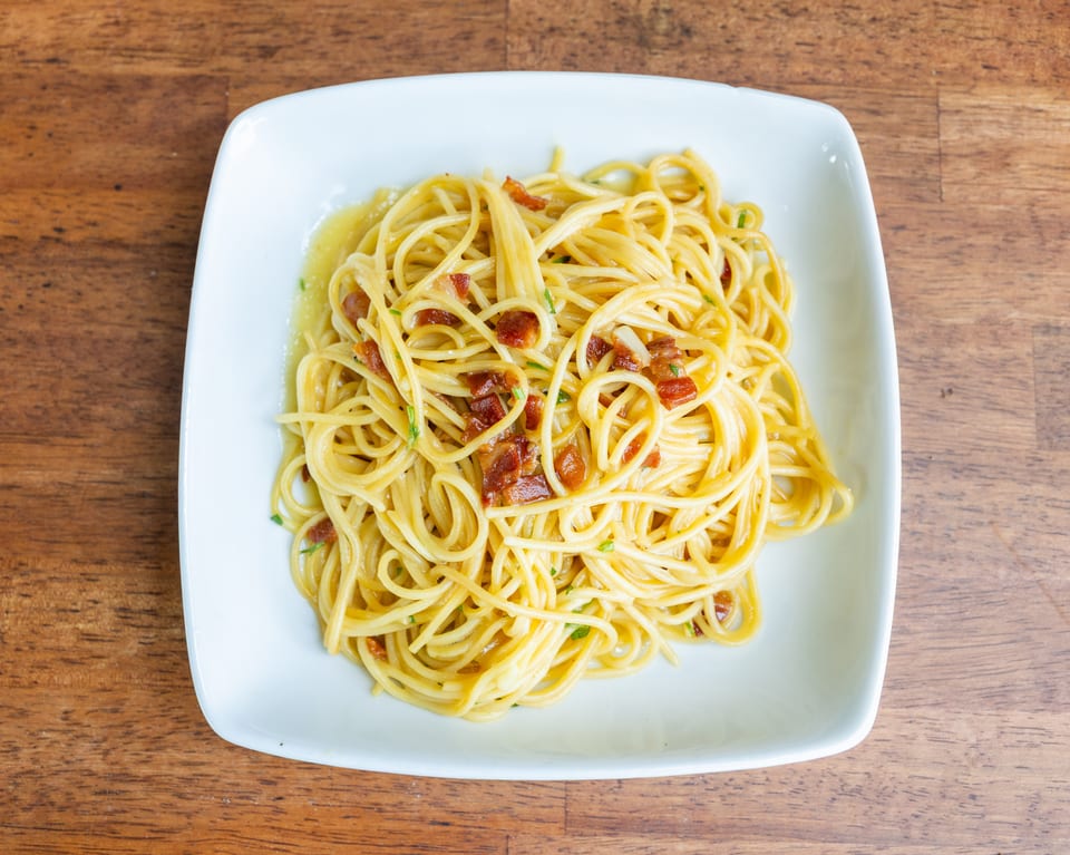 Spaghetti carbonara with crispy bacon bits and fresh herbs on a white square plate.