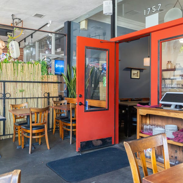 Restaurant entrance with an open red door, outdoor seating, and a bamboo privacy screen.