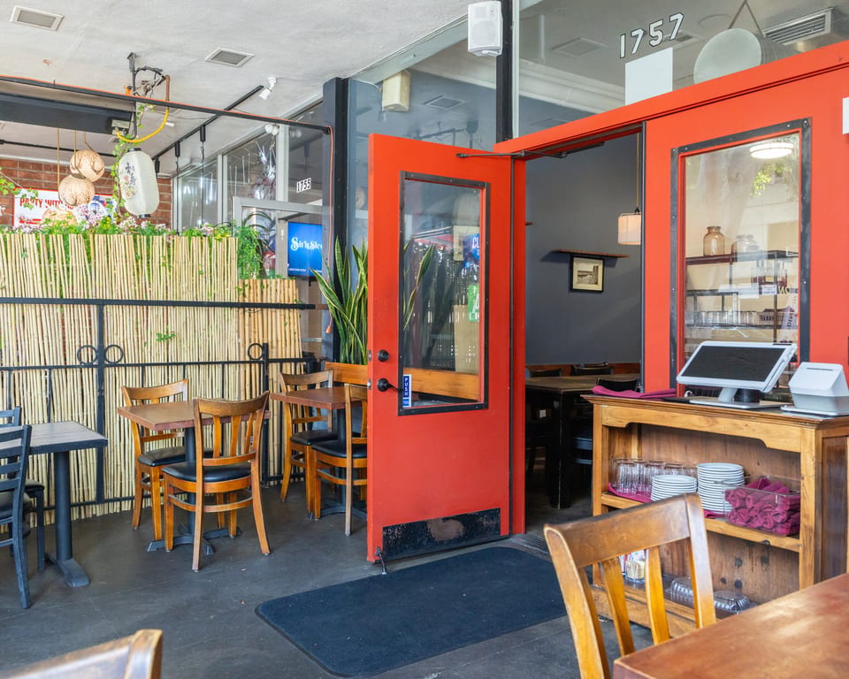 Restaurant entrance with an open red door, outdoor seating, and a bamboo privacy screen.