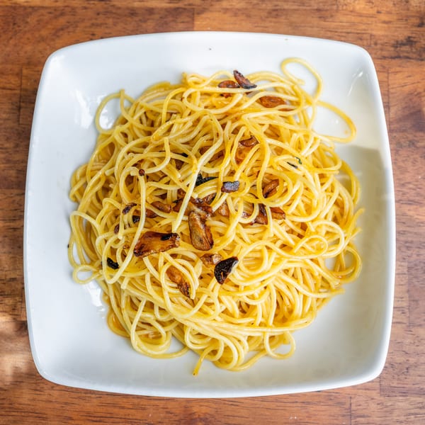 A white square plate filled with spaghetti aglio e olio, topped with crispy garlic slices, on a wooden table.