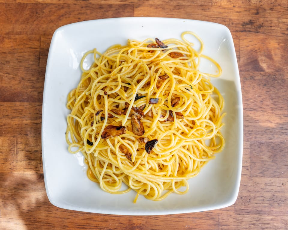 A white square plate filled with spaghetti aglio e olio, topped with crispy garlic slices, on a wooden table.
