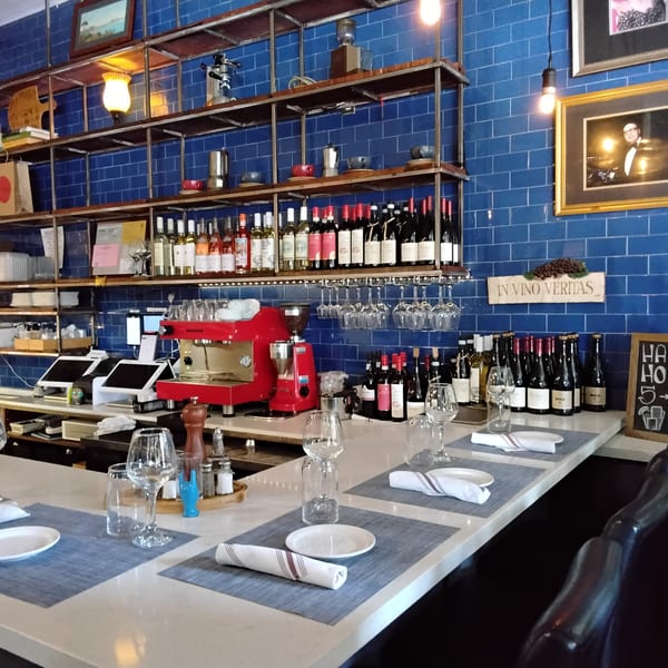 A restaurant bar with blue tiled walls, shelves of wine, and place settings on a white counter.