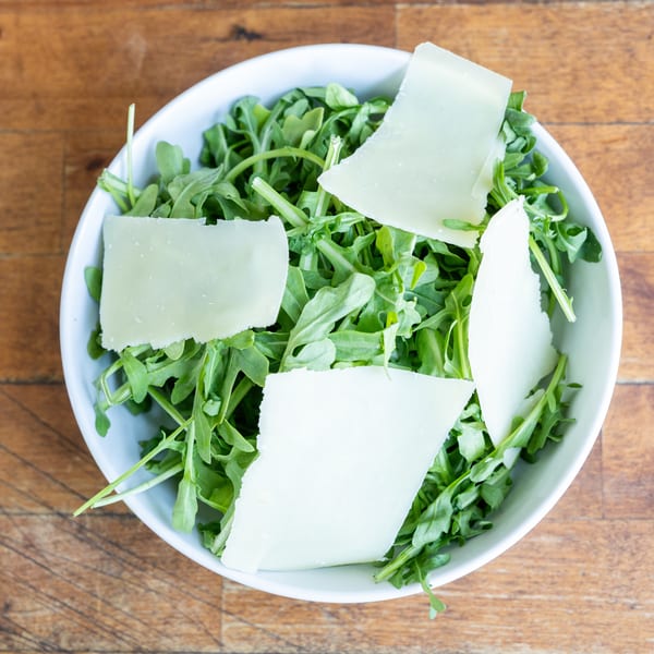 Overhead view of a white bowl filled with fresh arugula and large shavings of Parmesan cheese.
