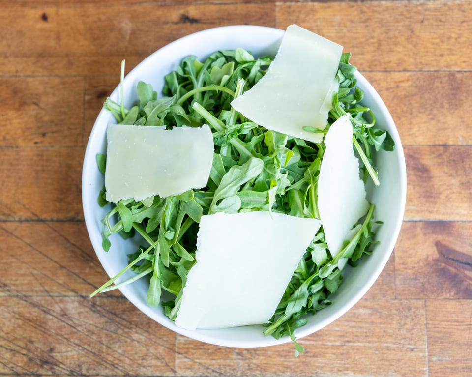 Overhead view of a white bowl filled with fresh arugula and large shavings of Parmesan cheese.