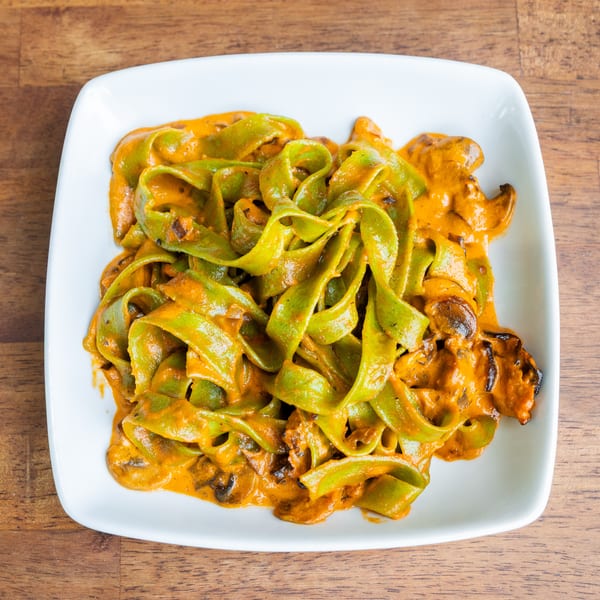 Green pappardelle pasta with a creamy orange sauce and mushrooms on a white plate.