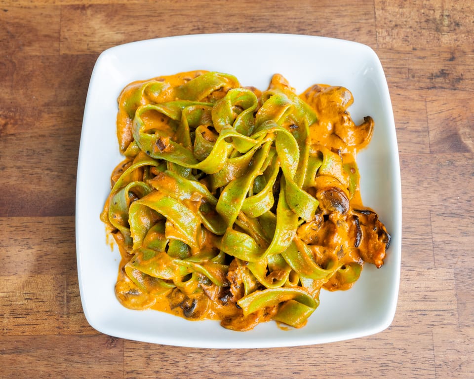 Green pappardelle pasta with a creamy orange sauce and mushrooms on a white plate.