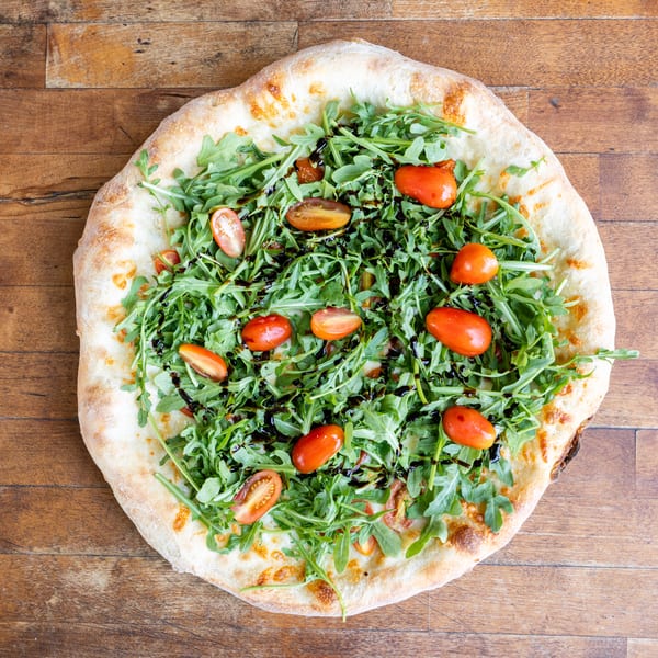 Overhead view of a pizza topped with fresh arugula, cherry tomatoes, and balsamic glaze on a wooden table.