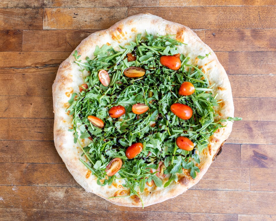 Overhead view of a pizza topped with fresh arugula, cherry tomatoes, and balsamic glaze on a wooden table.