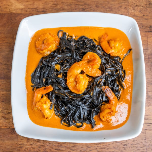 Black squid ink pasta with large orange shrimp in a vibrant red sauce on a white plate.