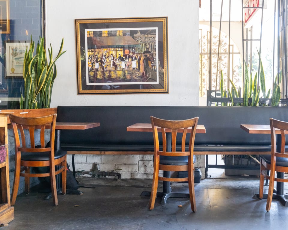 Restaurant interior with a long black booth, wooden tables and chairs, and a framed painting.