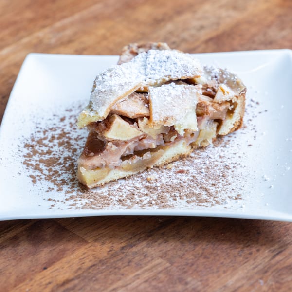 A slice of apple pie with a lattice crust, powdered sugar, and cinnamon on a white plate.