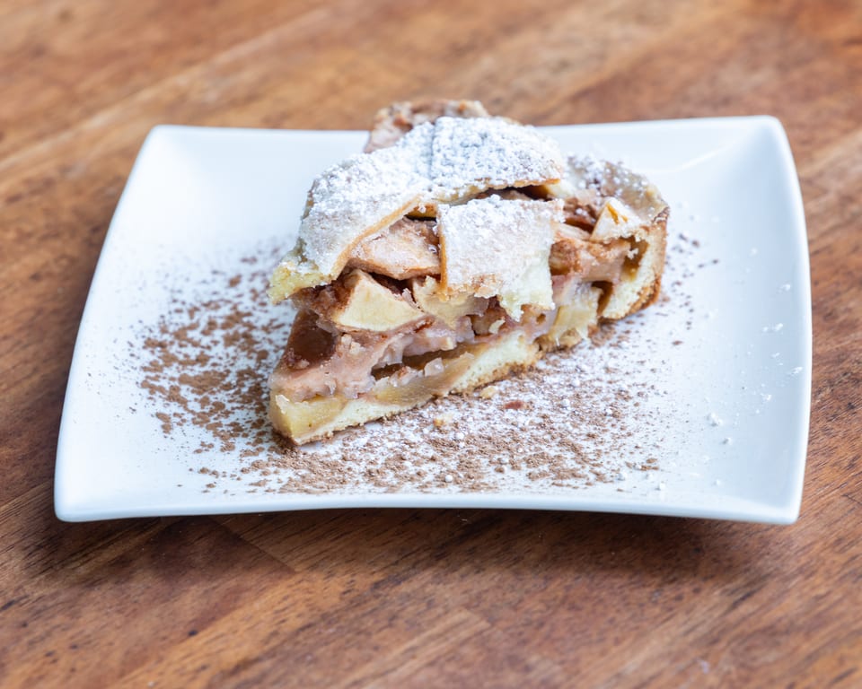 A slice of apple pie with a lattice crust, powdered sugar, and cinnamon on a white plate.