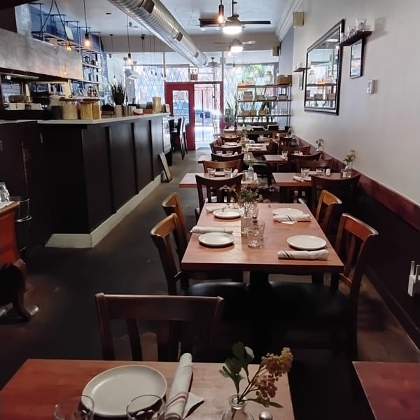 A long, narrow restaurant dining room with wooden tables, chairs, and a bar area.