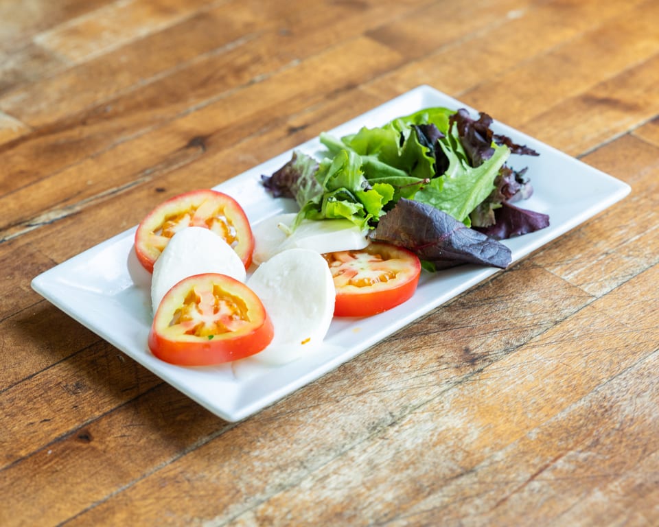 Caprese salad with sliced tomatoes, mozzarella, and mixed greens on a white rectangular plate.